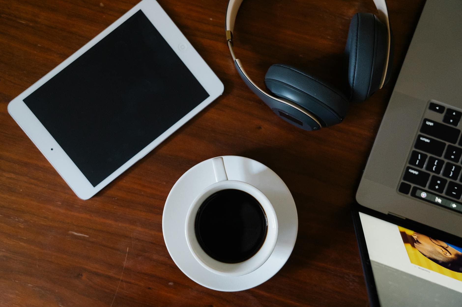 Flat lay of a coffee cup, tablet, headphones, and laptop for faceless YouTube creator workspace.